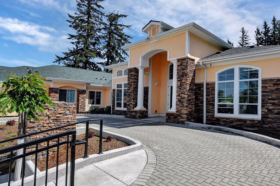 Exterior view of Tabor Crest Memory Care facility showing a peach-colored building with stone accents, large windows, and a covered entrance. The driveway and landscaped area with small plants and a tree are visible under a partly cloudy blue sky.