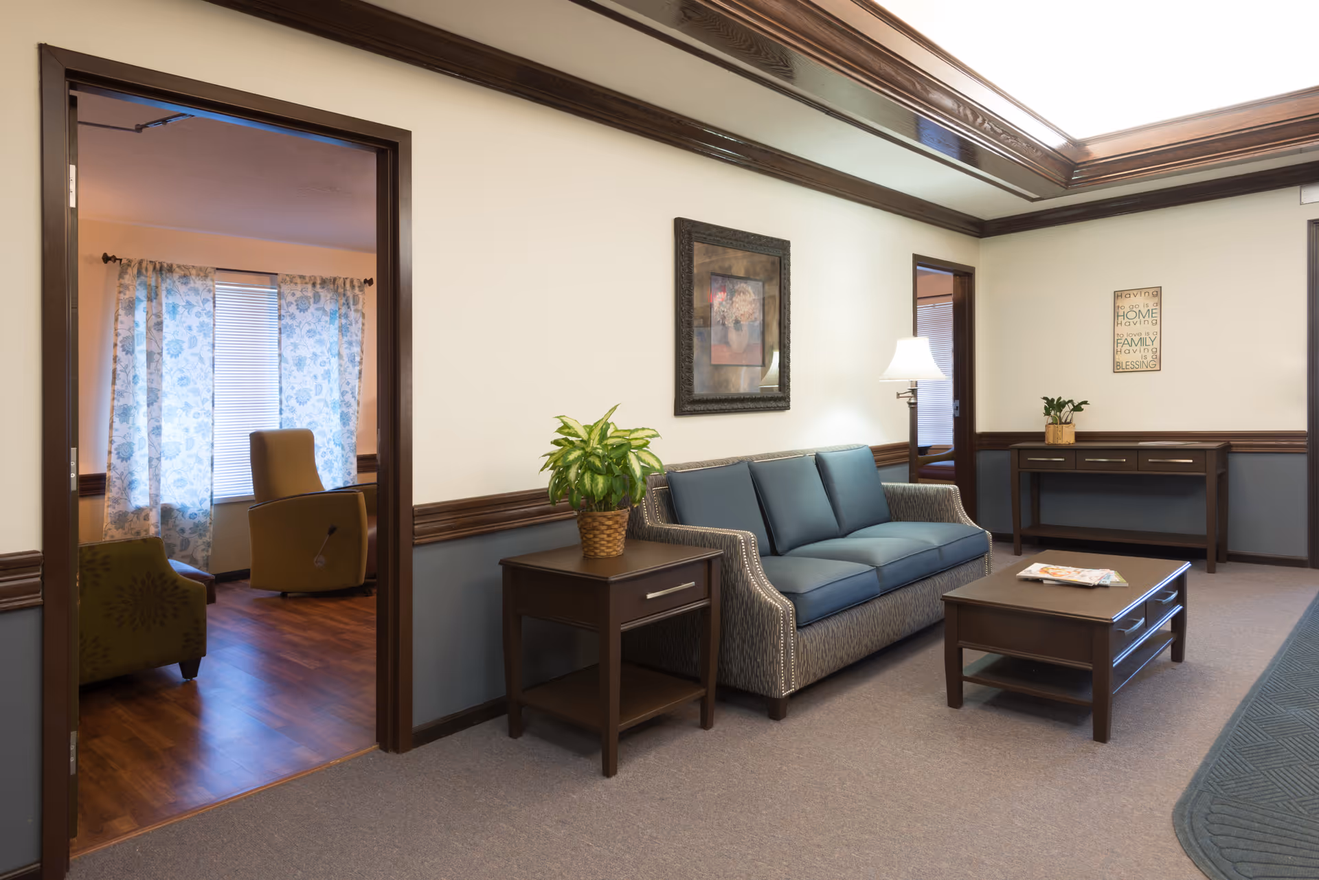 A cozy living room area in a senior living facility with a blue cushioned sofa, a wooden coffee table with magazines, a side table with a potted plant, and a floor lamp. The room has beige walls with dark wood trim and a framed picture hanging above the sofa. Through an open doorway, another room with two armchairs and floral curtains on the window is visible.