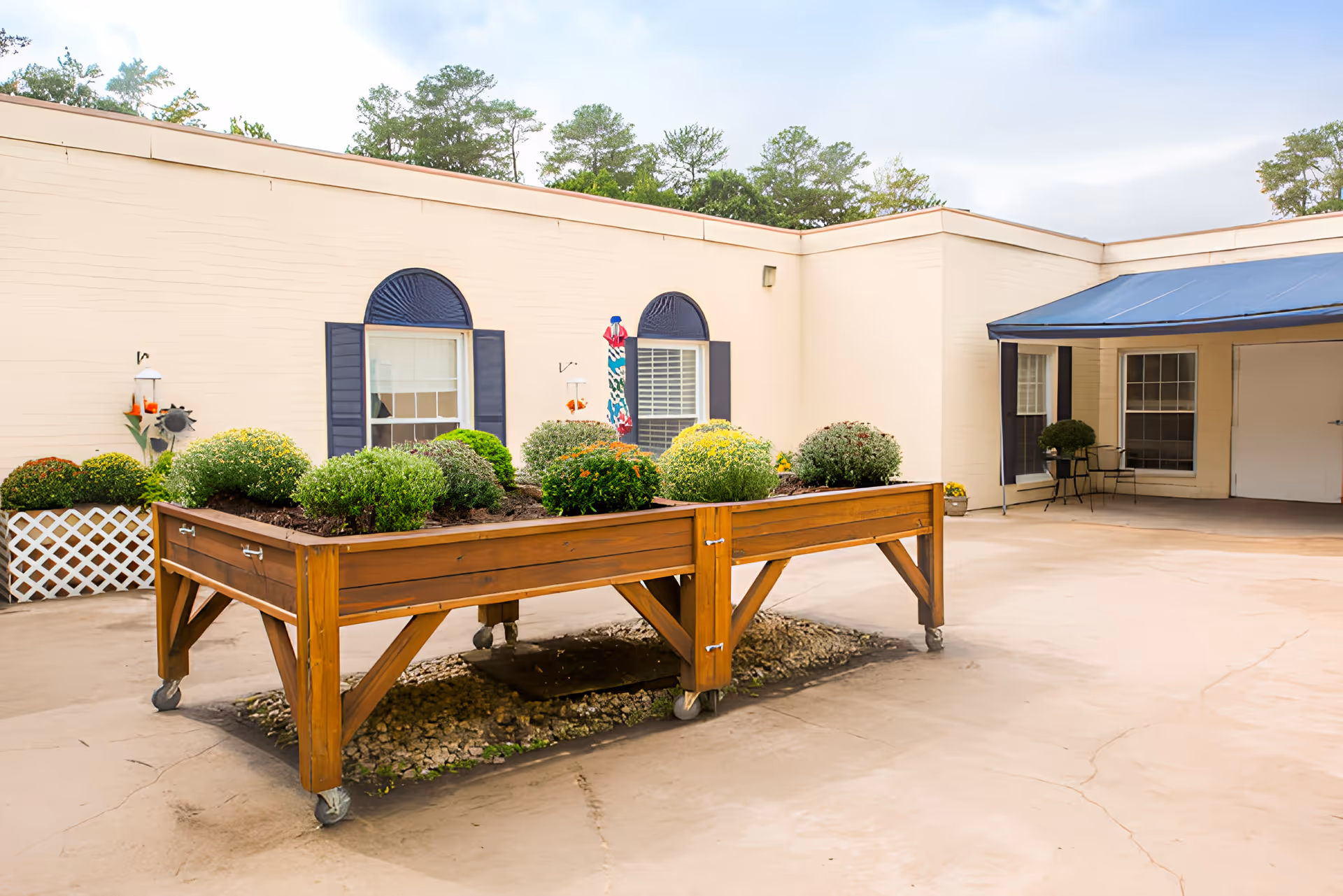 Outdoor courtyard with a large raised wooden planter on casters filled with shrubs in front of a beige single-story building and covered entry.