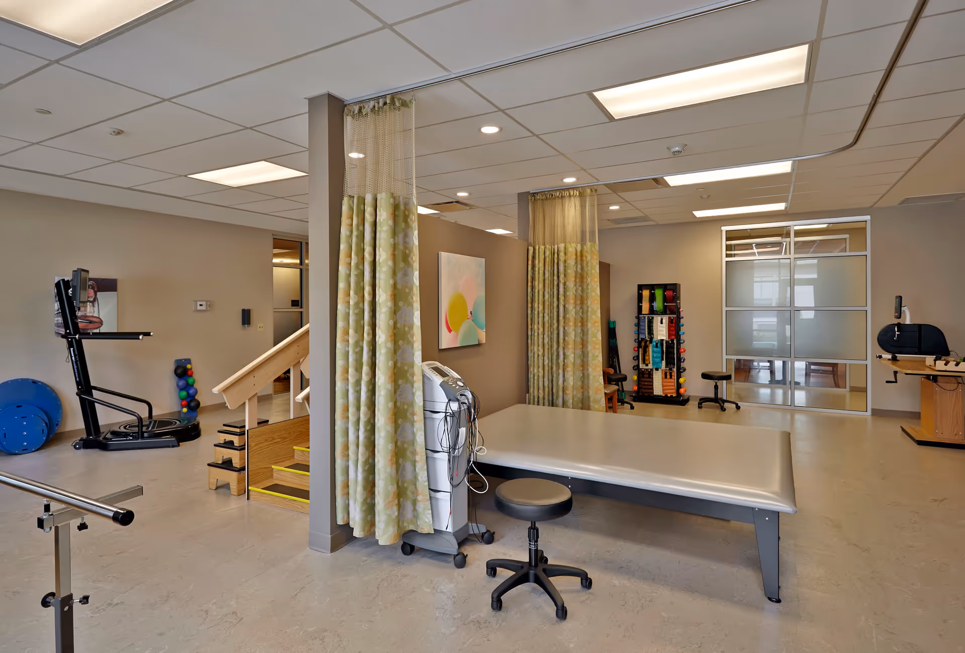 A bright physical therapy room with a treatment table, rolling stool, exercise equipment, therapy stairs, and privacy curtains.