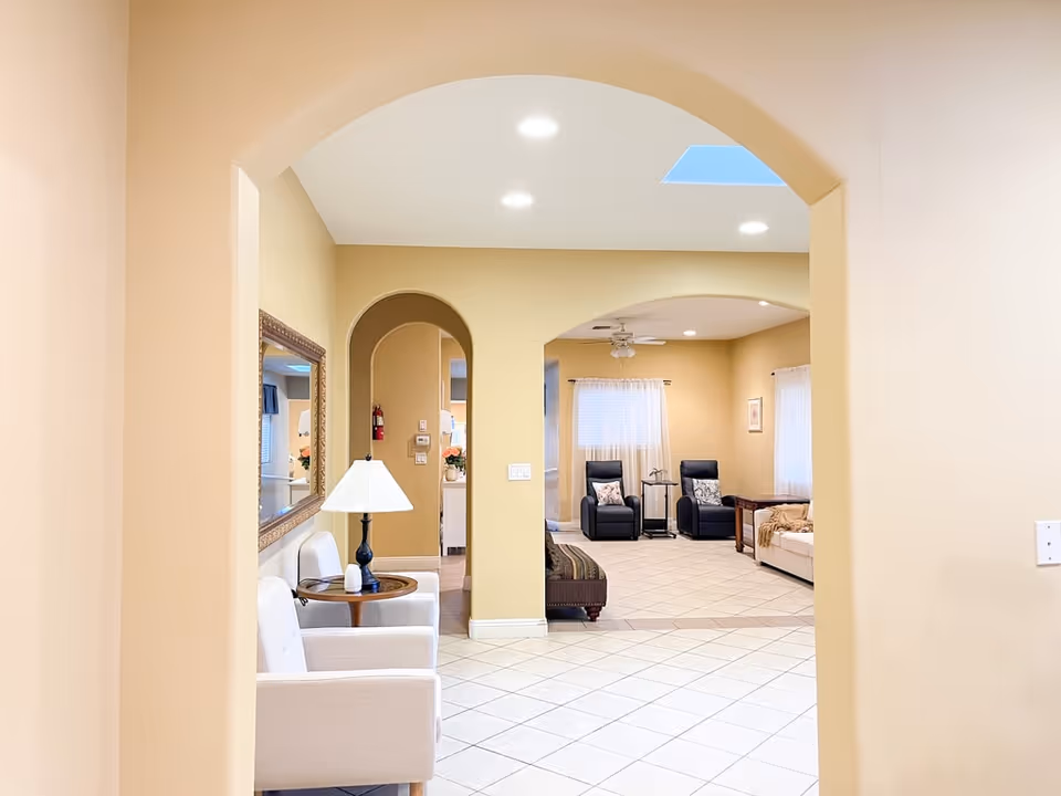 Interior view of a senior living facility common area with beige walls and tiled floors. The space features two beige armchairs with a small round wooden table and lamp between them on the left side. In the background, there are two black recliners with pillows, a small table between them, and a beige sofa with a throw blanket. The room is well-lit with recessed ceiling lights and natural light from windows with sheer curtains. There are arched doorways and a ceiling fan in the background.