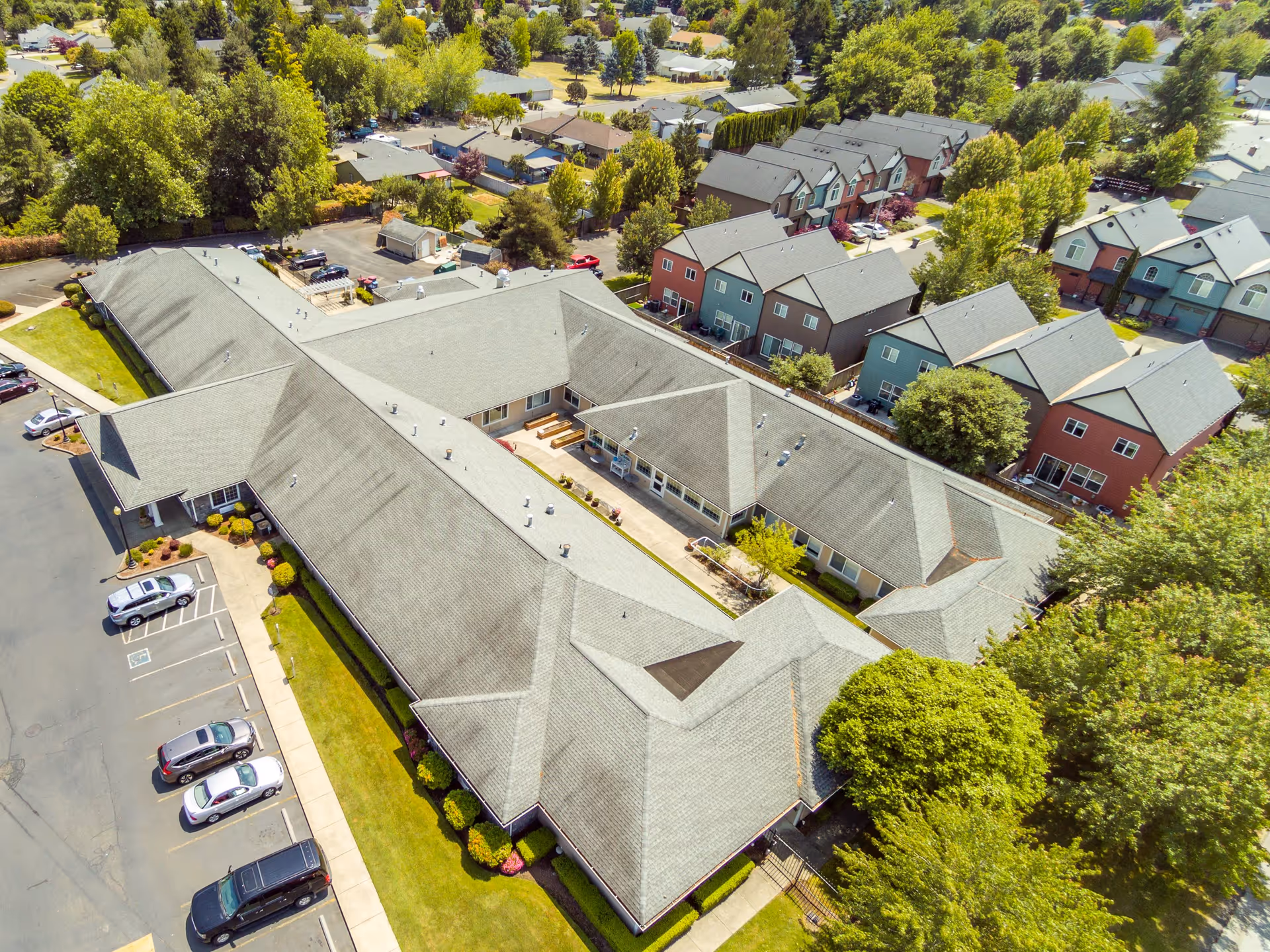 Aerial view of McMinnville Memory Care facility showing a large, single-story building with a gray roof surrounded by trees and a parking lot with several cars. Residential houses and greenery are visible in the background.