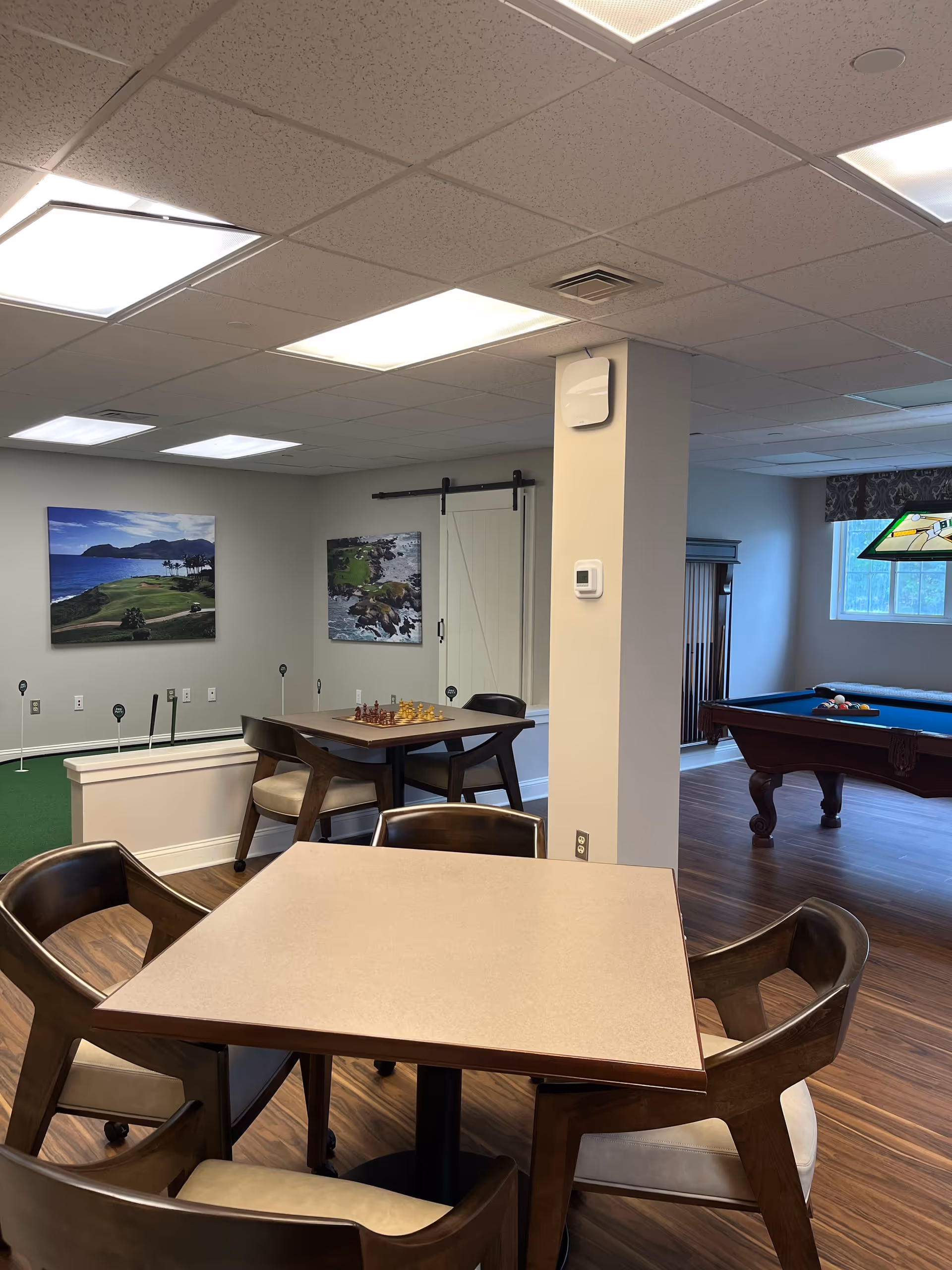Interior view of a recreational room in a senior living facility featuring a square table with four chairs in the foreground, a smaller table with a chess set, a putting green area with golf holes, and a pool table near a window with a stained glass light fixture above it. The room has wood flooring, neutral-colored walls, and ceiling lights.