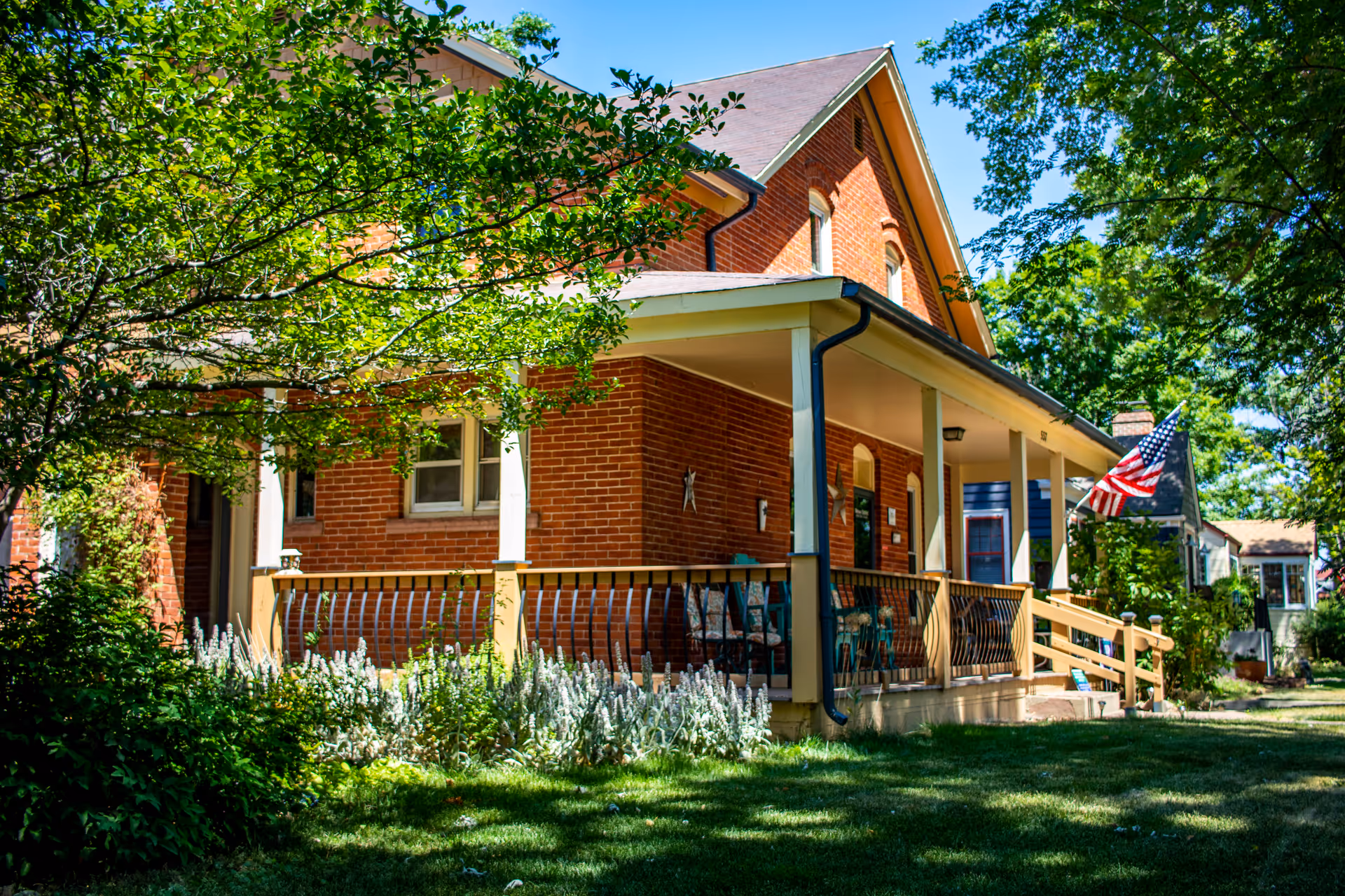 A red brick house with a covered porch featuring several chairs and a railing. The porch has white pillars and a ramp leading up to it. There is an American flag displayed near the porch. The house is surrounded by green trees, bushes, and a well-maintained lawn under a clear blue sky.