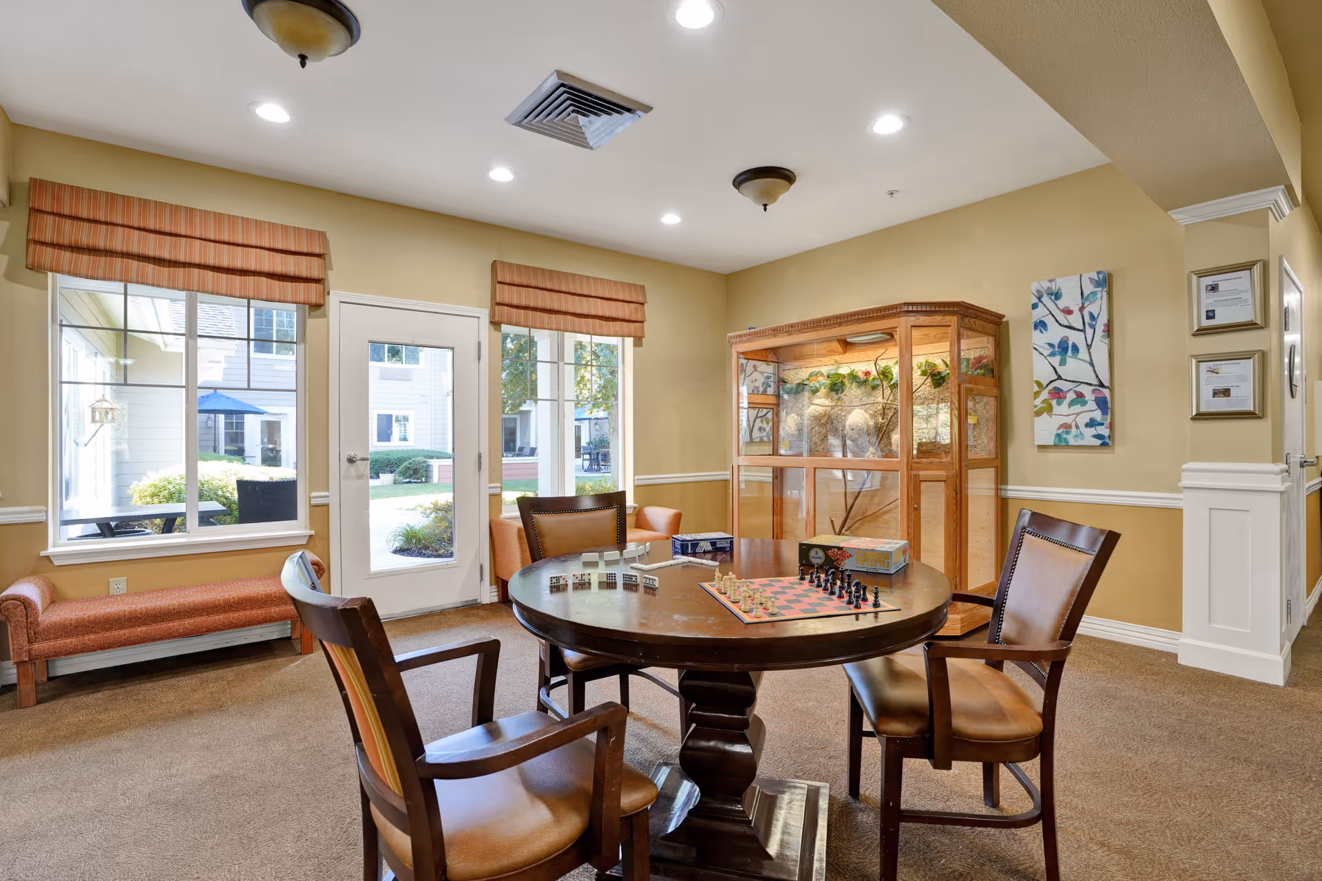 A cozy common area in a senior living facility with a round wooden table set for chess and other board games, surrounded by four wooden chairs with leather seats. The room has beige walls, carpeted floor, two large windows and a glass door leading outside, a cushioned bench under one window, and a large wooden display cabinet with plants inside. Wall art and framed certificates decorate the walls.
