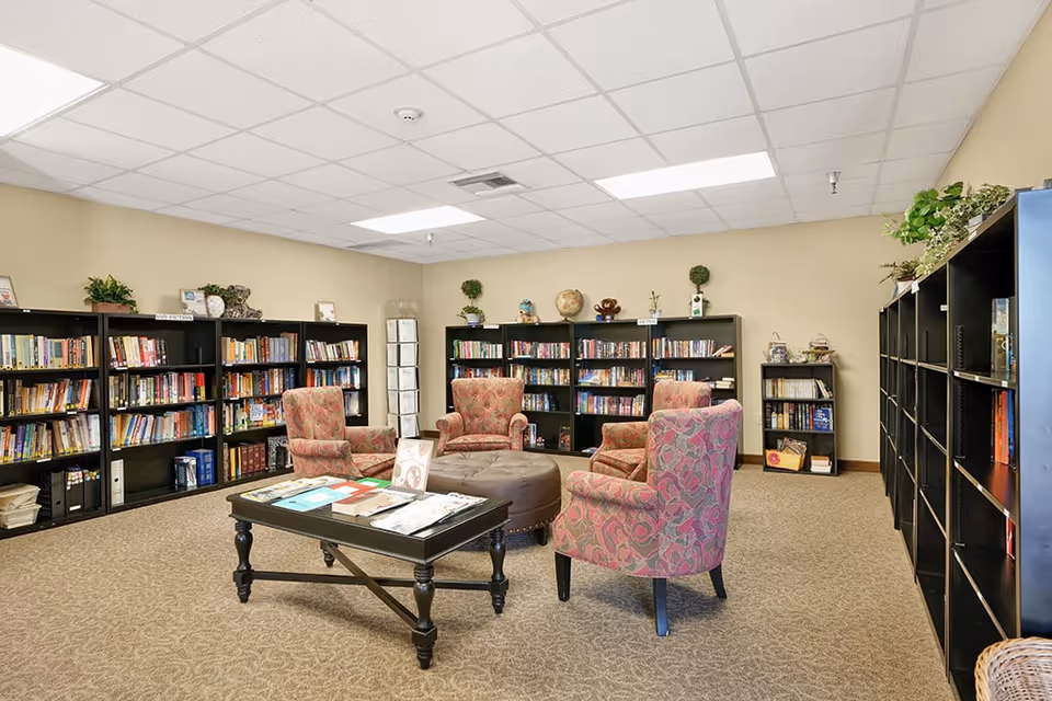 A cozy library room with beige walls and carpeted floor, featuring multiple black bookshelves filled with books and decorative items. Four patterned armchairs are arranged around a round ottoman and a rectangular coffee table with magazines and books on it. The ceiling has white tiles with fluorescent lighting.