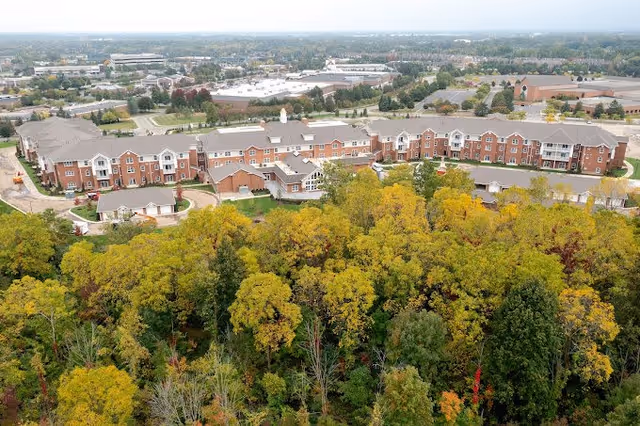 Aerial view of Willow Pines Retirement Community showing a large, multi-story brick building surrounded by trees with autumn foliage. The facility is situated near a suburban area with other buildings and parking lots visible in the background.