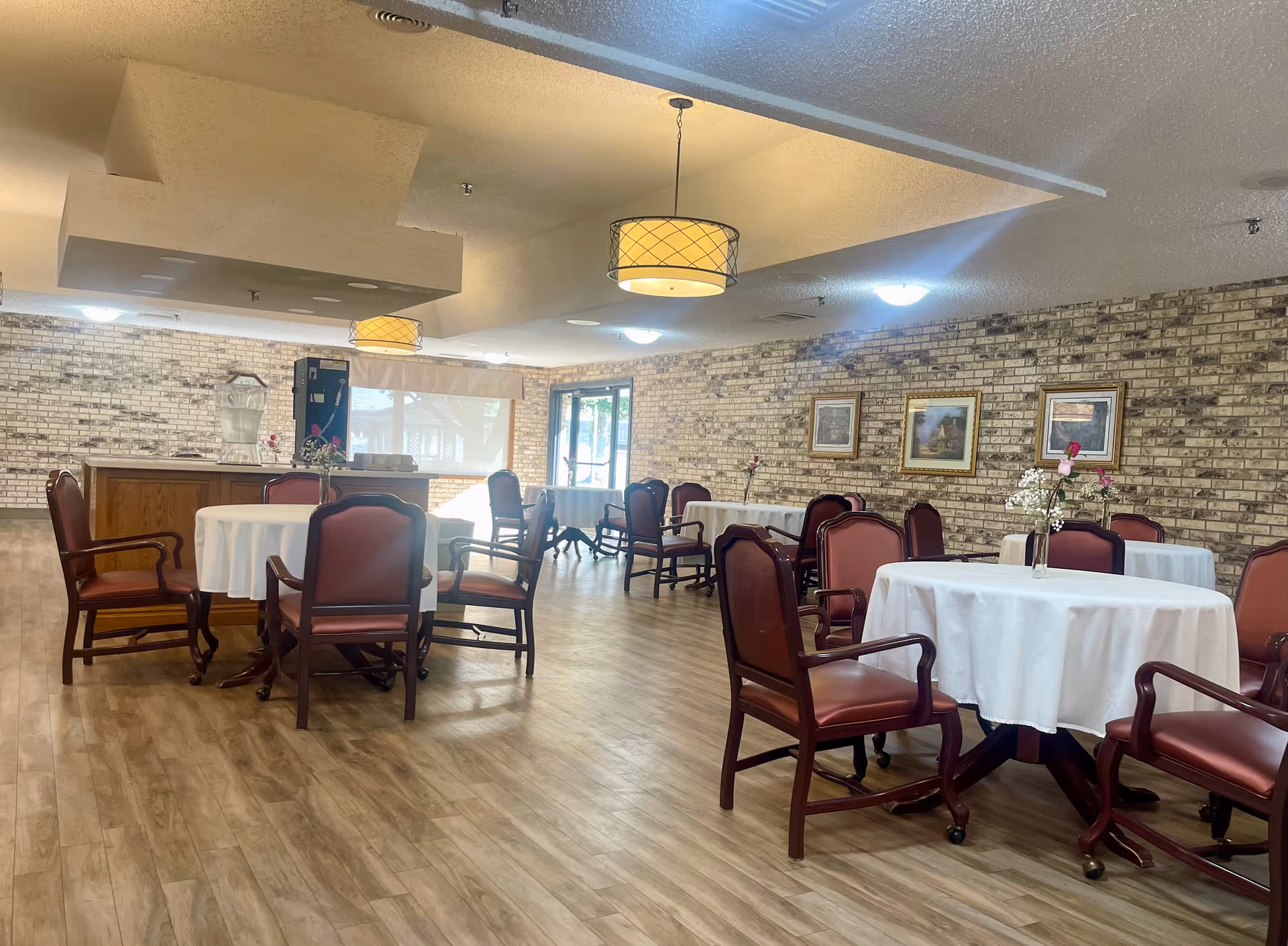 A dining room in a senior living facility with round tables covered in white tablecloths, surrounded by wooden chairs with red cushions. The room has wood flooring, brick walls adorned with framed paintings, and ceiling lights with decorative shades. A counter with a water dispenser and cups is visible in the background near a window.