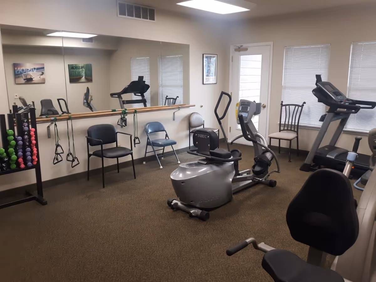 A small fitness room with exercise equipment including a recumbent bike, treadmill, and weights. There are several chairs along the walls, a large mirror, and resistance bands hanging from a wooden bar. The room has beige walls, carpeted floor, and windows with blinds.