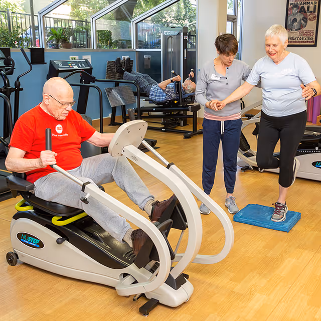An elderly man exercises on a seated fitness machine in a gym room while another elderly man uses a leg press machine in the background. A fitness instructor assists an elderly woman balancing on a blue foam pad. The room has large windows and wooden flooring.