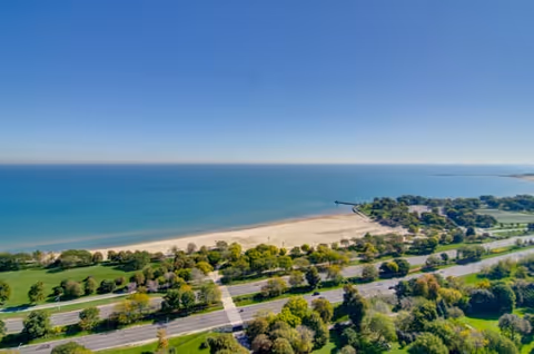 Aerial view of a beach with a wide sandy shore, calm blue water, and a clear sky. There is a green park area with many trees and a road running parallel to the beach.