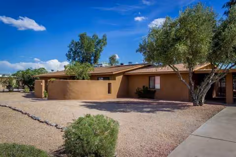 Exterior view of Sunrise Care Home Shea, a desert-style single-story facility with trees and gravel landscaping.