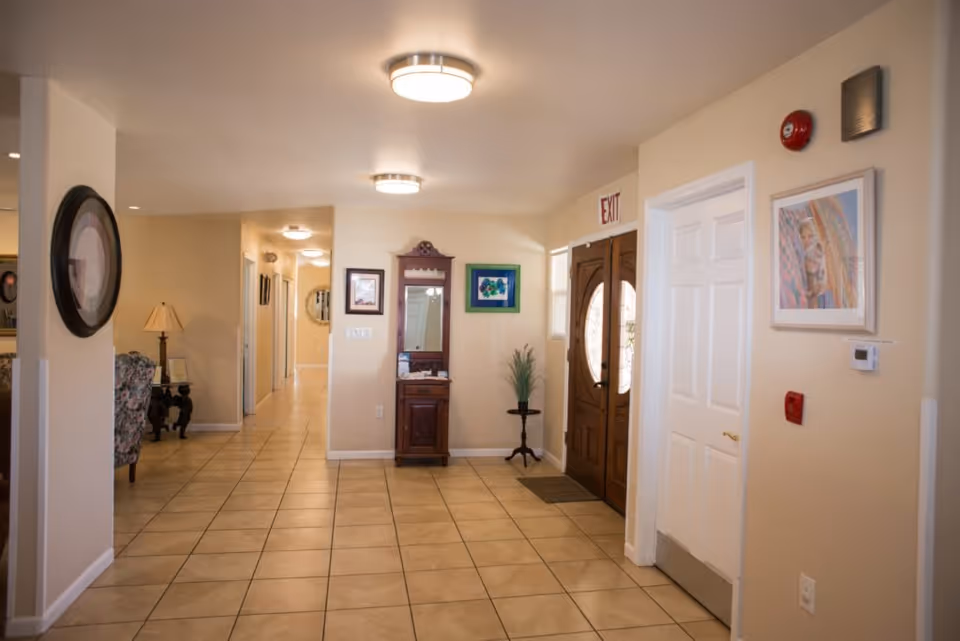 Interior hallway of an assisted living facility with beige tiled floor and cream-colored walls. There are wooden double doors with glass panels on the right side, a small wooden cabinet with a mirror above it, a framed picture, and a potted plant on a small stand. Ceiling lights illuminate the hallway, and there is an exit sign above the doors. A hallway extends further back with additional doors and a lamp on a side table visible on the left.