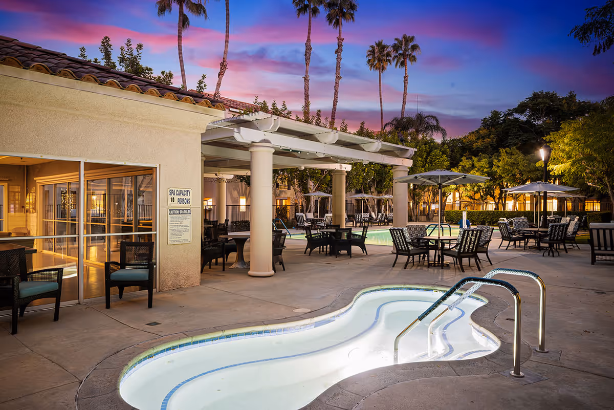 Outdoor pool and spa patio with tables, chairs, umbrellas, and palm trees under a colorful sunset sky.