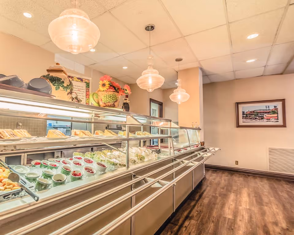 Interior view of a dining area at Tower Grove Manor featuring a buffet counter with various food items including sandwiches and bowls of fruit and salad. The room has hanging pendant lights, wooden flooring, and a framed picture on the wall.
