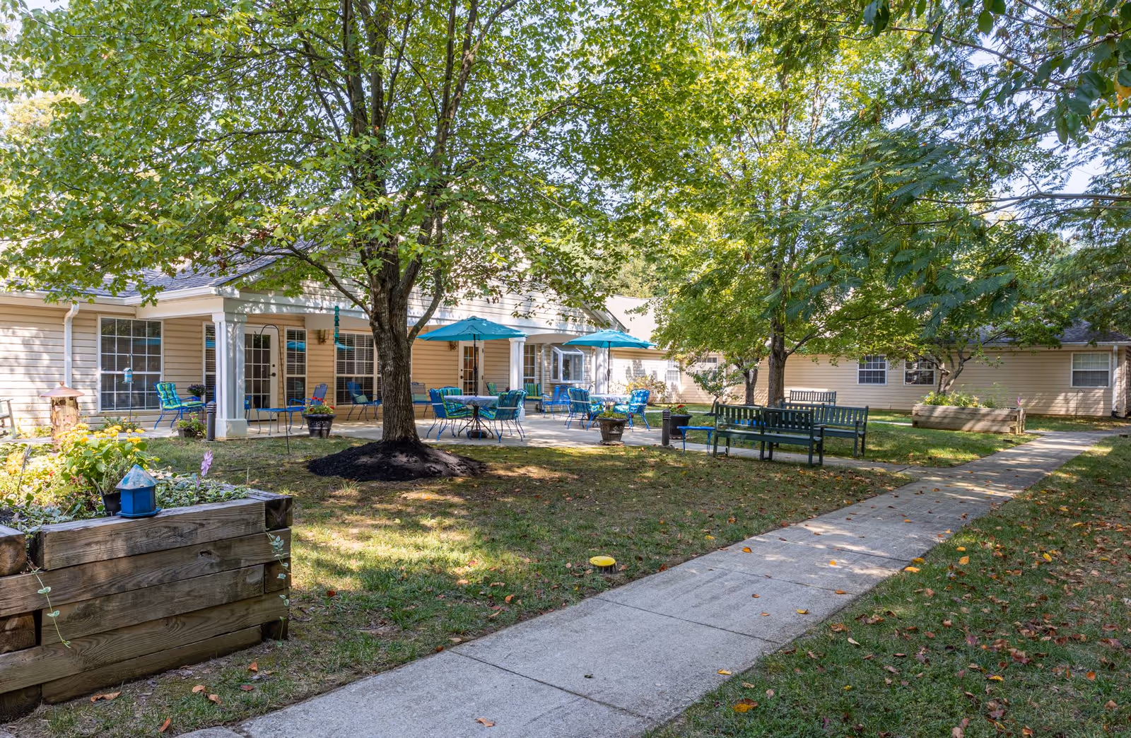 Outdoor patio area at Charter Senior Living of Annapolis with several blue metal tables and chairs under turquoise umbrellas. The patio is surrounded by trees and greenery, with a concrete walkway leading through the grassy area. There are wooden planter boxes with flowers and benches nearby.