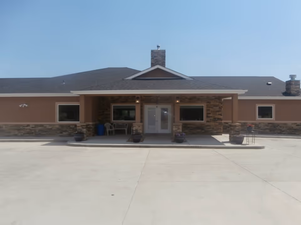 Front exterior view of a single-story assisted living facility building with a covered entrance, stone and brown siding, several windows, and a concrete driveway in front.
