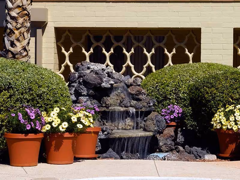 Small tiered rock water fountain with cascading water in front of a building facade, flanked by potted flowers and trimmed shrubs.