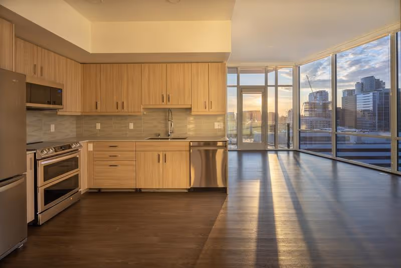 Modern kitchen area with light wood cabinets, stainless steel appliances including a refrigerator, oven, microwave, and dishwasher. The kitchen opens into a spacious living area with large floor-to-ceiling windows offering a view of a city skyline at sunset.