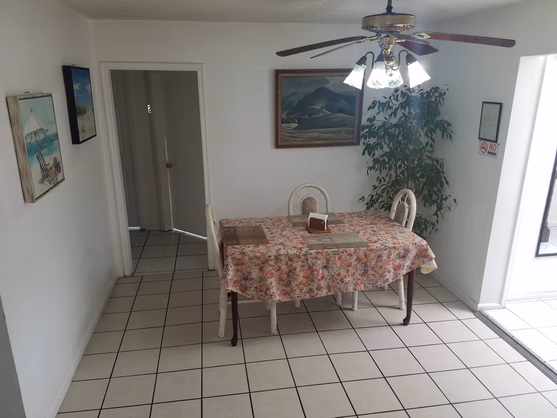 A small dining area with a rectangular table covered with a floral tablecloth and four chairs. Above the table is a ceiling fan with lights. On the wall behind the table is a framed landscape painting and a large potted plant. The floor is tiled with white tiles, and there are two paintings on the left wall. A 'No Smoking' sign is visible on the right wall near a doorway.