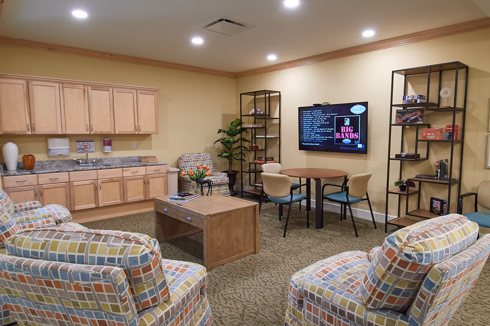 A cozy senior living common room with patterned armchairs, a coffee table, kitchenette cabinets, a round table with chairs, shelving, and a wall-mounted TV.