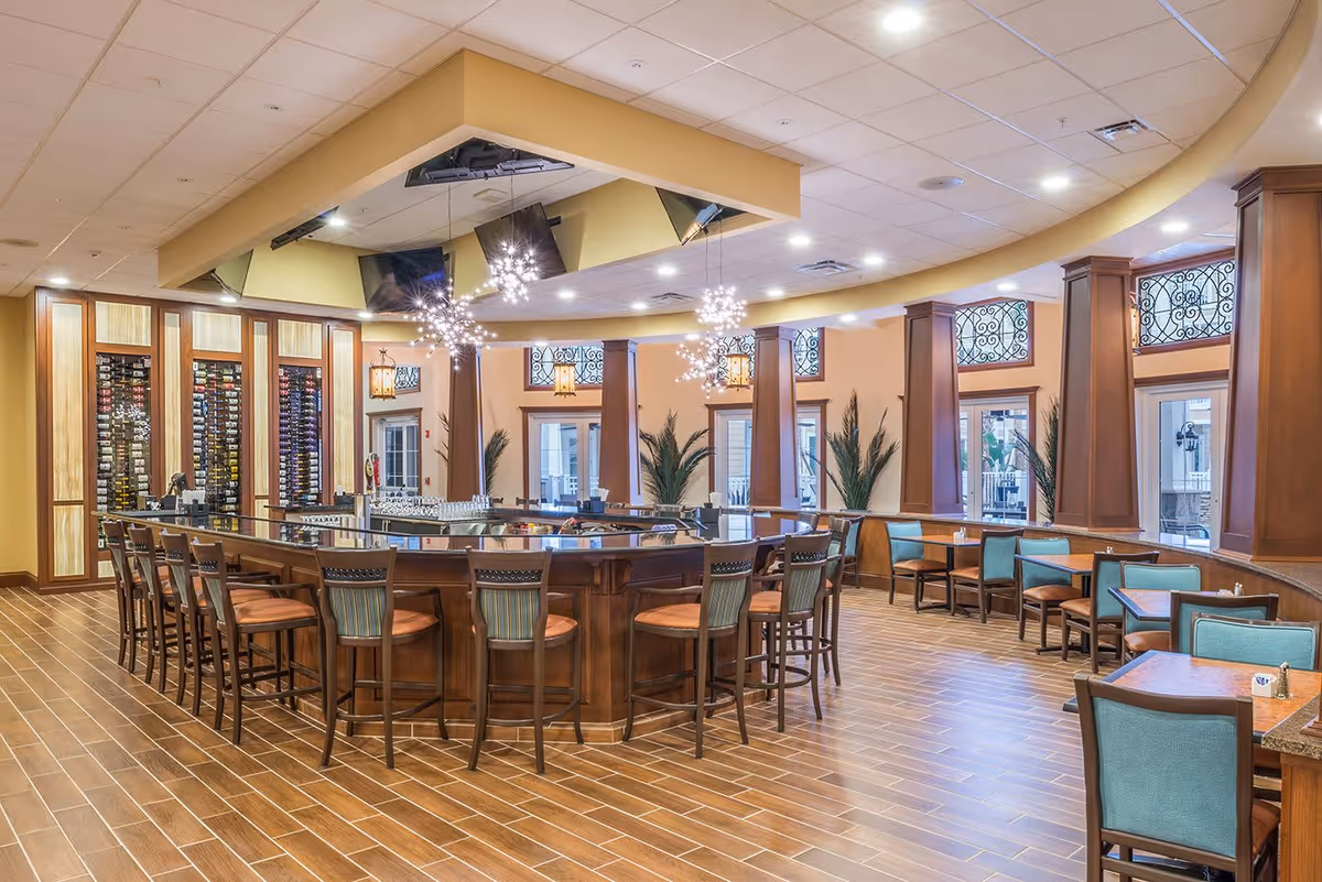 A spacious and well-lit dining area with a large central bar surrounded by wooden bar stools. The room features warm yellow walls, decorative light fixtures, and several tables with teal upholstered chairs along the windows. There are tall windows with decorative ironwork and plants placed between the columns.