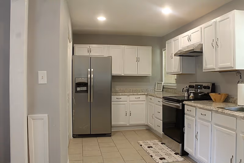A modern kitchen with white cabinets, granite countertops, a stainless steel refrigerator, stove, and range hood. The floor is tiled, and there is a small window with blinds above the counter. A patterned rug is placed in front of the stove.