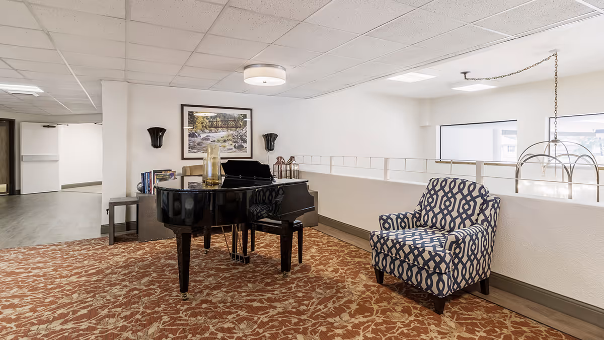 Interior common area with a black grand piano, a patterned armchair, a small table with books and decorative items, a framed picture on the wall, and ceiling lights. The area has a patterned carpet and white walls with large windows providing natural light.