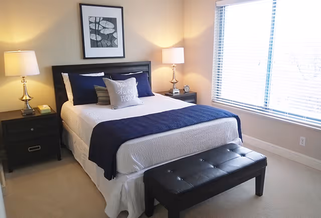 A neatly made bed with white bedding and a navy blue throw blanket in a bedroom. The bed has several pillows, including one with the text 'Home Sweet Home'. There are two bedside tables with lamps on either side of the bed. A black cushioned bench is placed at the foot of the bed. A framed black and white artwork hangs above the headboard. A large window with blinds allows natural light into the room.