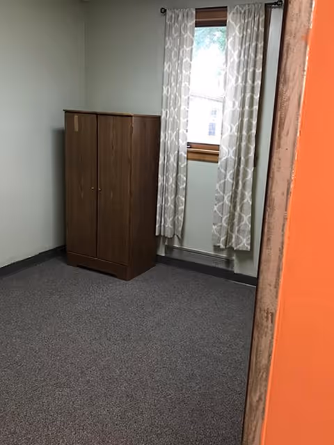 A small, sparsely furnished room with gray carpet, light green walls, a wooden wardrobe against the wall, and a window with patterned curtains letting in natural light.
