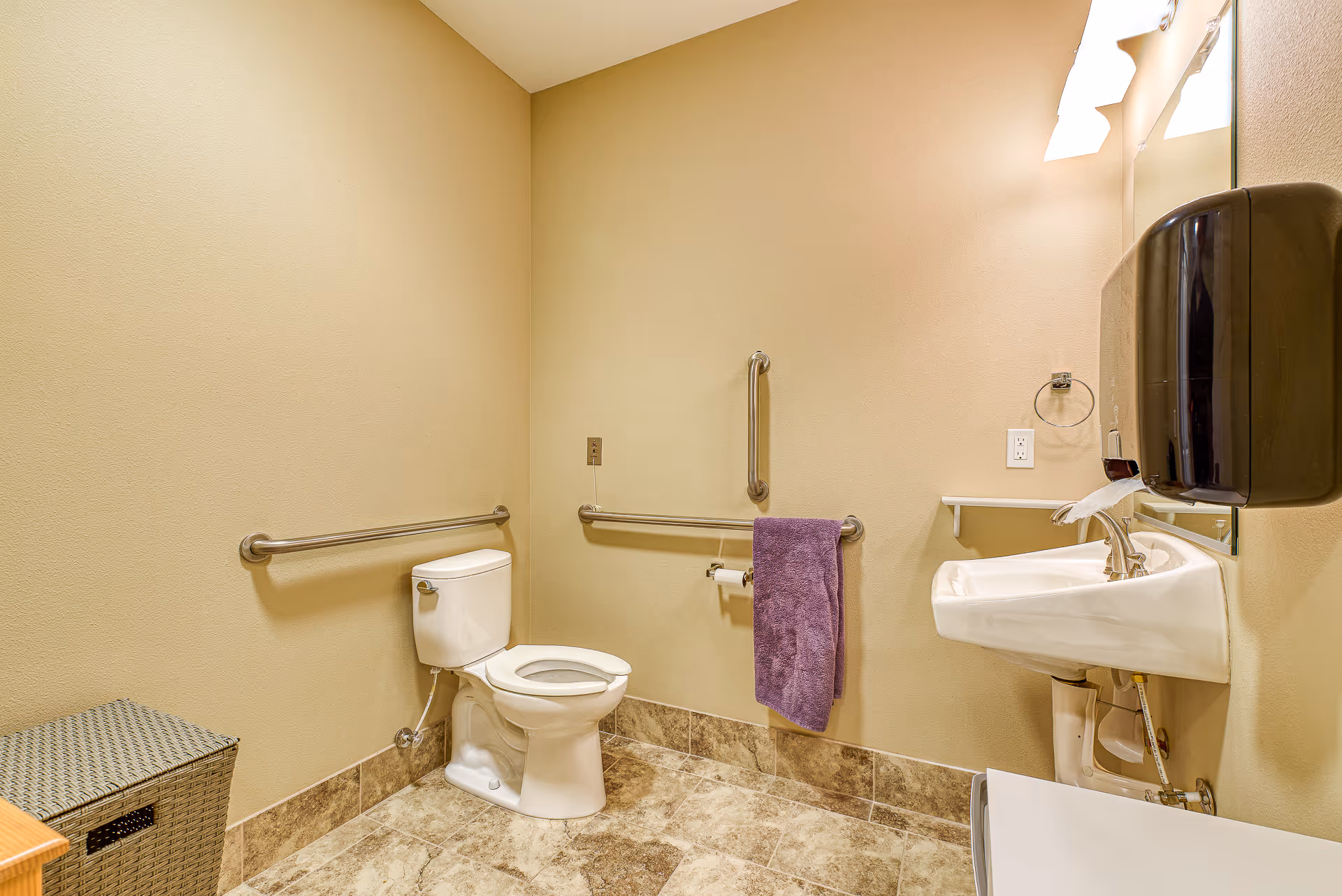 A clean, accessible bathroom with beige walls and tiled floor. It features a white toilet with grab bars on the walls around it, a white sink with a mirror above, a black soap dispenser mounted on the wall, a purple towel hanging on a grab bar, and a woven laundry basket in the corner.