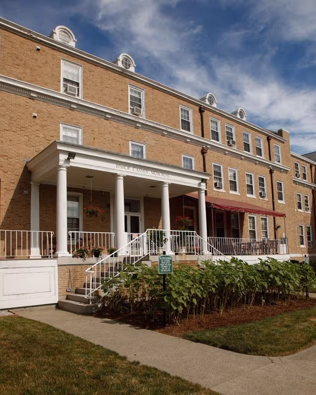 Brick three-story skilled nursing building with white columns at the main entrance, a raised porch, and landscaped flowerbeds.