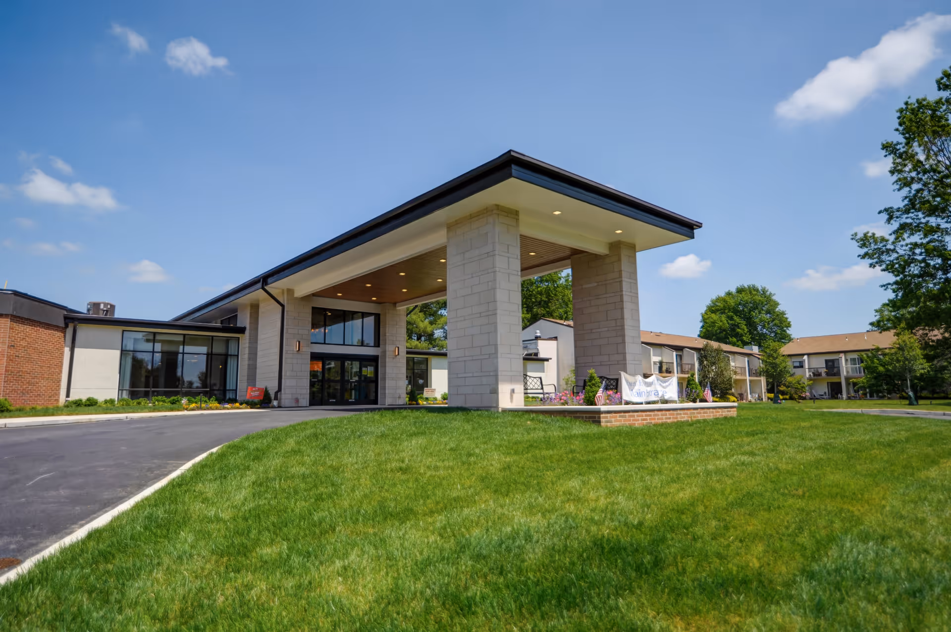 Exterior view of Wesley Enhanced Living Main Line facility showing a modern entrance with large pillars and a covered driveway. The building is surrounded by a well-maintained green lawn under a blue sky with scattered clouds.
