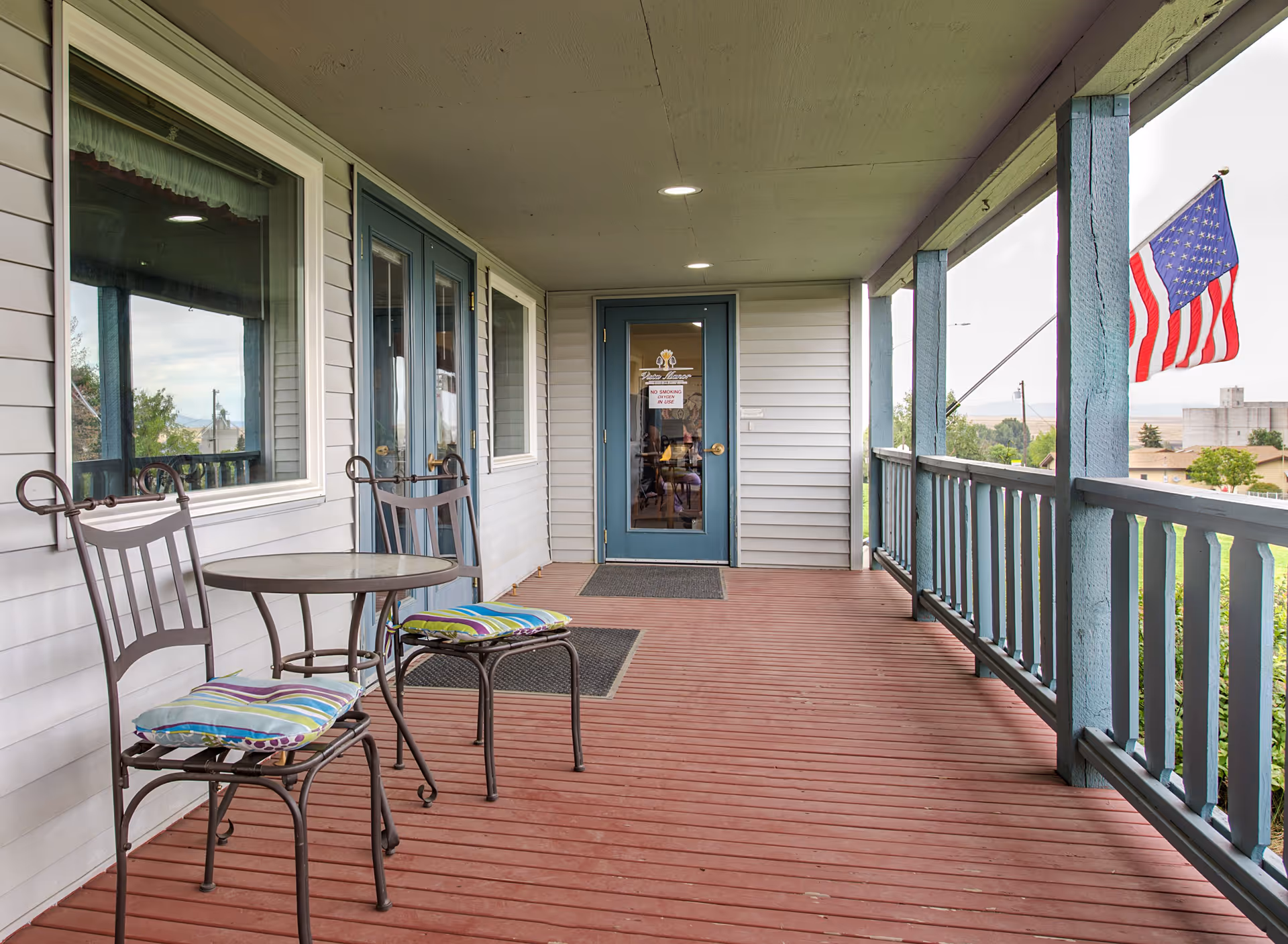 Covered porch area with red wooden flooring, two metal chairs with colorful cushions, and a small round table. The porch has blue railings and posts, with a door and windows leading inside the building. An American flag is visible on the right side, and some greenery and buildings can be seen in the background.