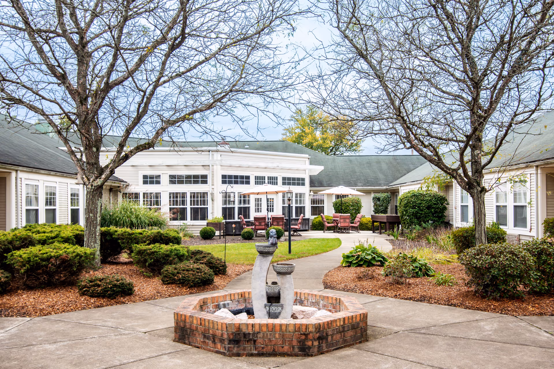 Courtyard with a brick fountain, landscaped beds, trees, and patio seating in front of a single-story senior living building.