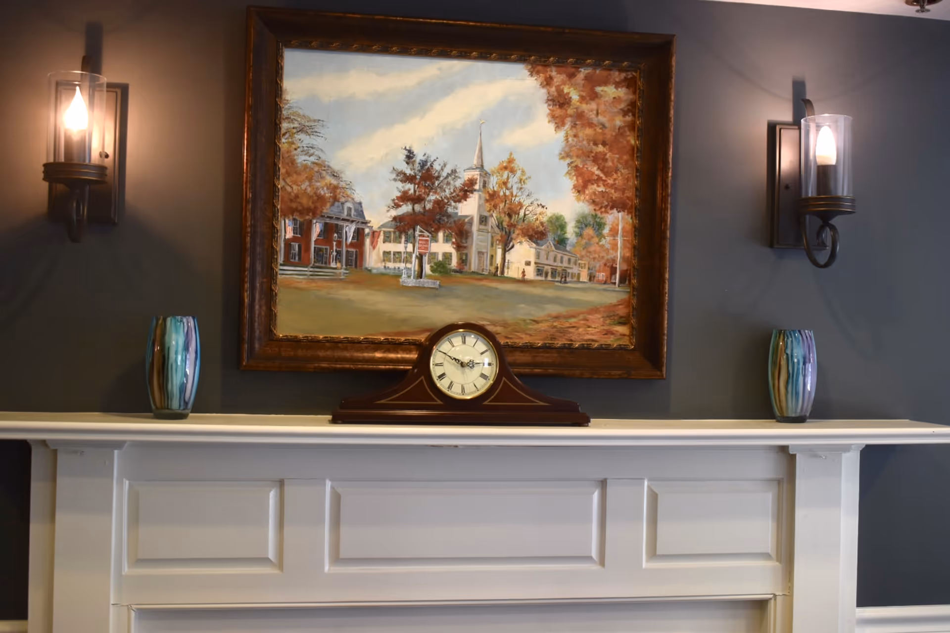 A white mantelpiece with a wooden clock in the center and two decorative vases on either side. Above the mantel hangs a framed painting depicting a scenic view of buildings and trees with autumn foliage. Two wall-mounted candle-style light fixtures flank the painting on a gray wall.