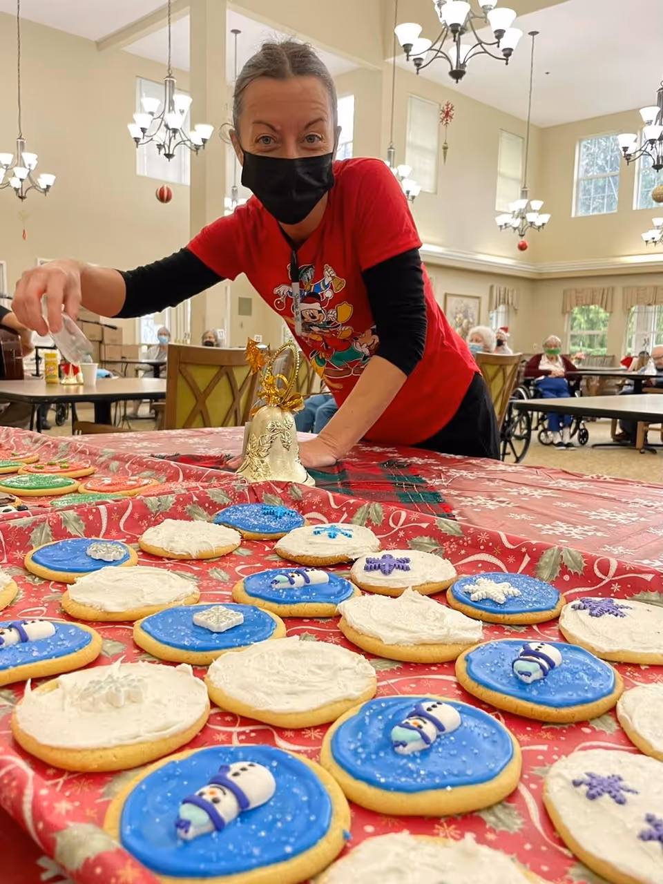 A woman wearing a black face mask and a red holiday-themed shirt decorates Christmas cookies on a table covered with a red festive tablecloth. The cookies are decorated with blue and white icing, featuring snowmen and snowflakes. In the background, elderly people are seated in a spacious, well-lit room with large windows and chandeliers.
