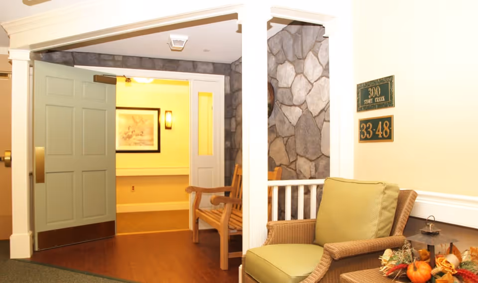A cozy indoor seating area with a green cushioned wicker chair and a wooden bench near an open green door leading to a hallway with framed artwork on the wall. The walls have a stone accent and there are signs indicating room numbers 300 and 33-48.