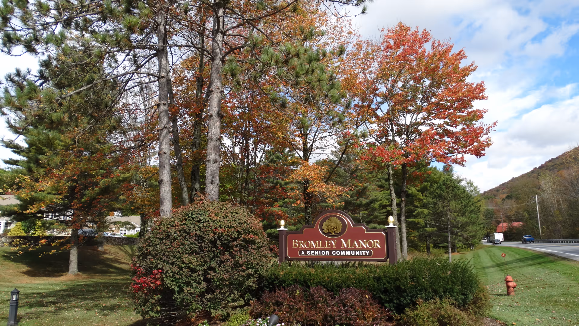Outdoor view of the entrance sign for Bromley Manor, a senior community, surrounded by trees with autumn foliage and green bushes, next to a road with a fire hydrant and a few vehicles in the distance.