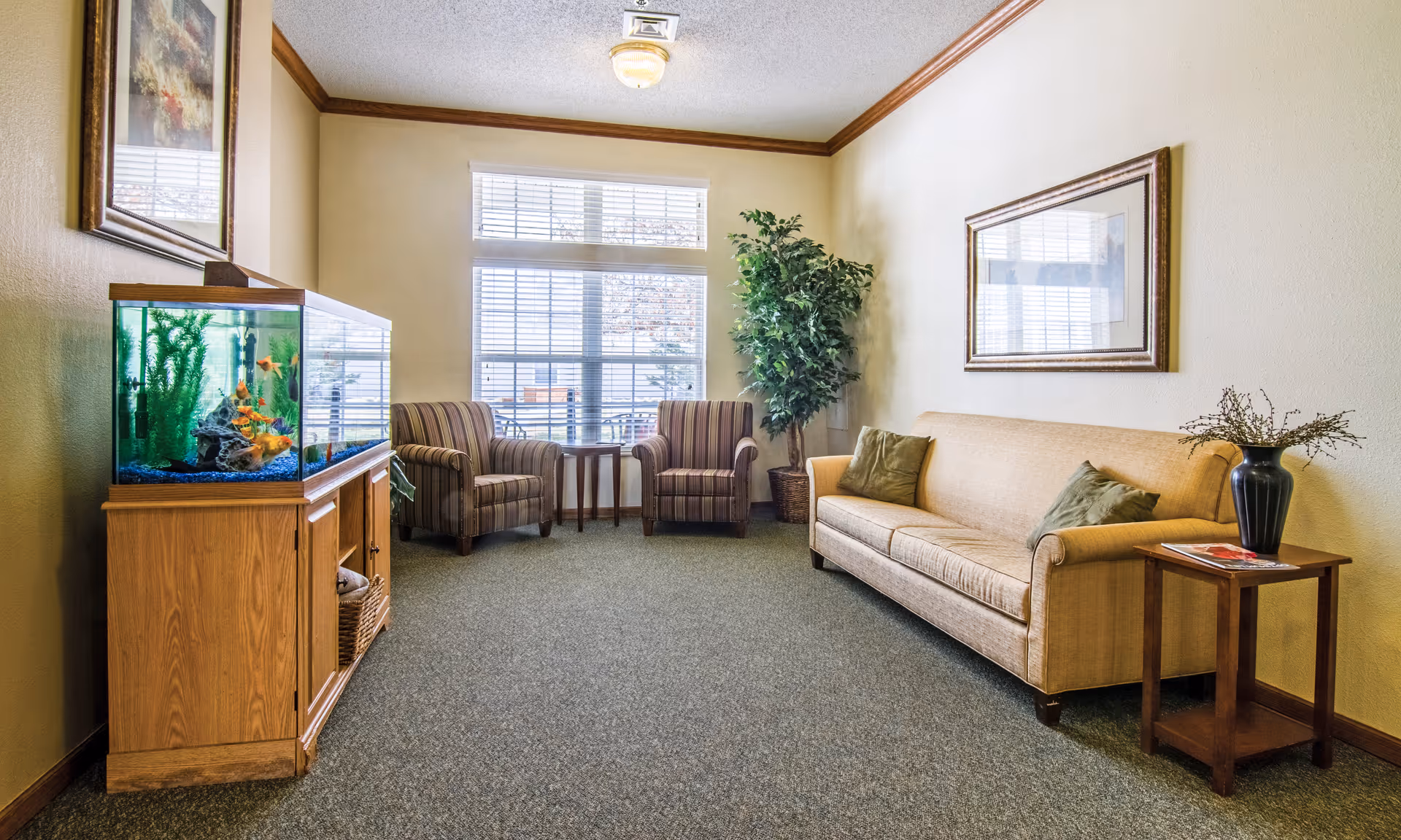 Well-lit sitting area with a sofa, two armchairs, side tables, a large fish tank, and a potted plant in front of a window.