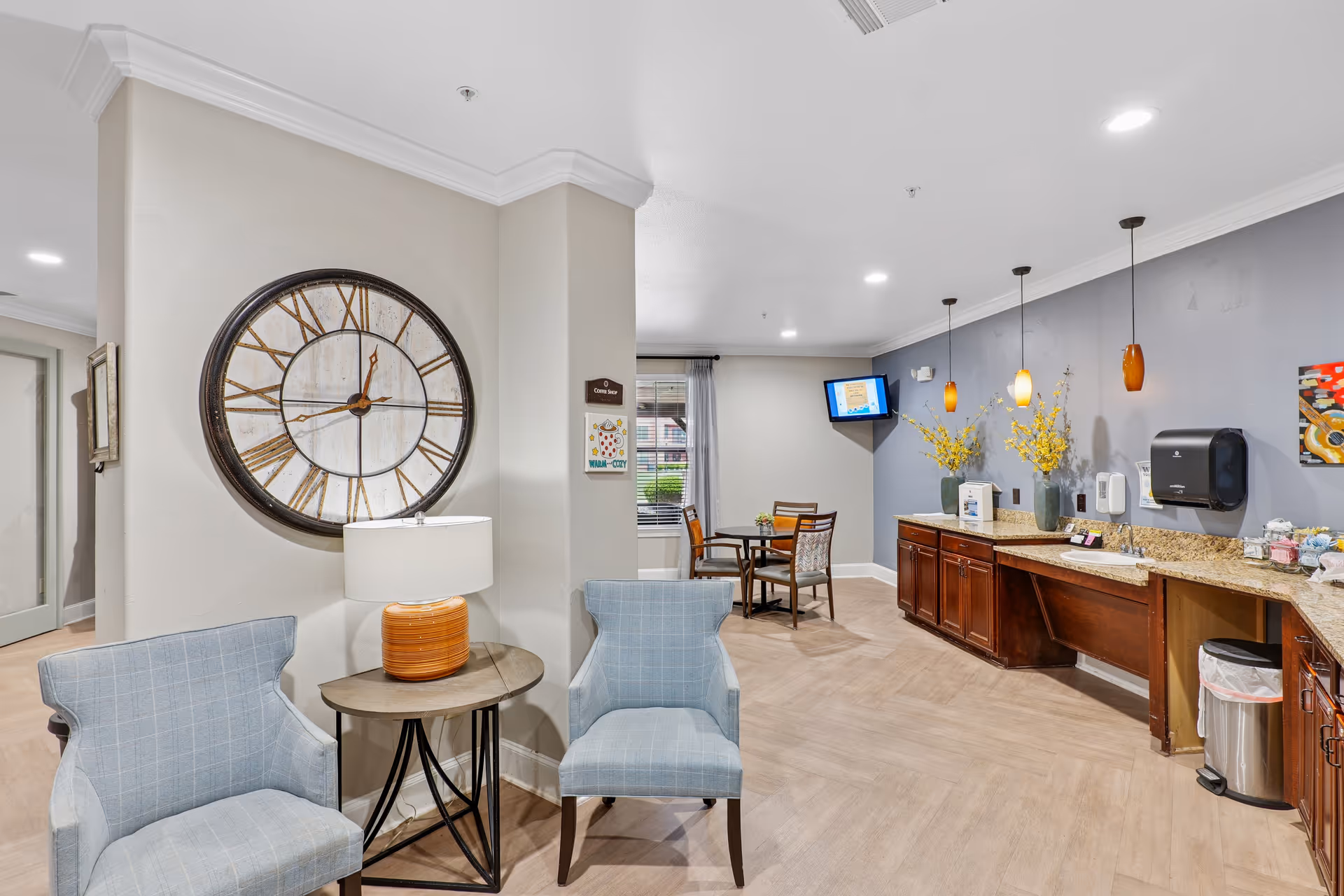 A cozy interior common area with two light blue armchairs and a small round table with an orange lamp. A large decorative wall clock hangs on the wall behind the chairs. In the background, there is a small dining table with chairs near a window, a wall-mounted TV, and a long countertop with cabinets, a sink, and decorative yellow flowers in vases. The walls are painted light gray and blue, and the floor has a light wood pattern.