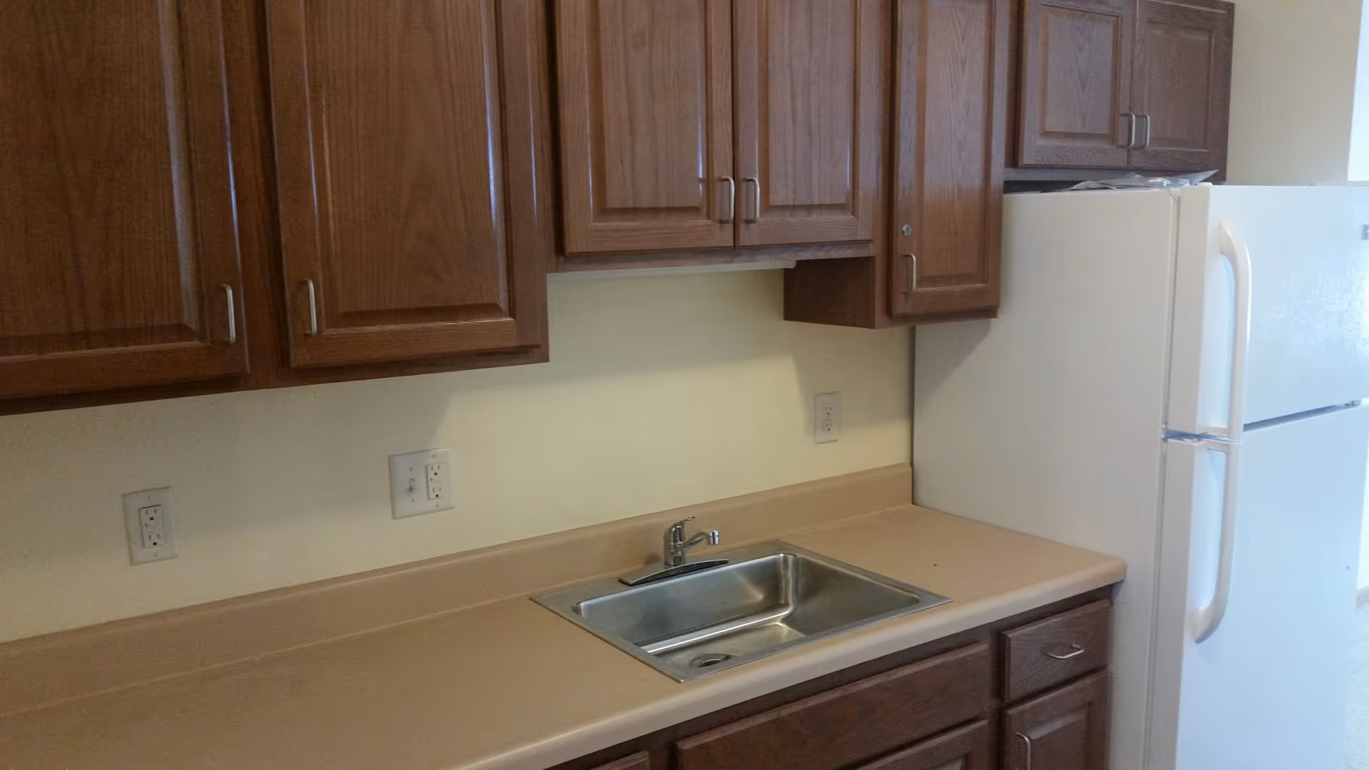 A kitchen area with wooden cabinets, a beige countertop, a stainless steel sink with a faucet, and a white refrigerator.