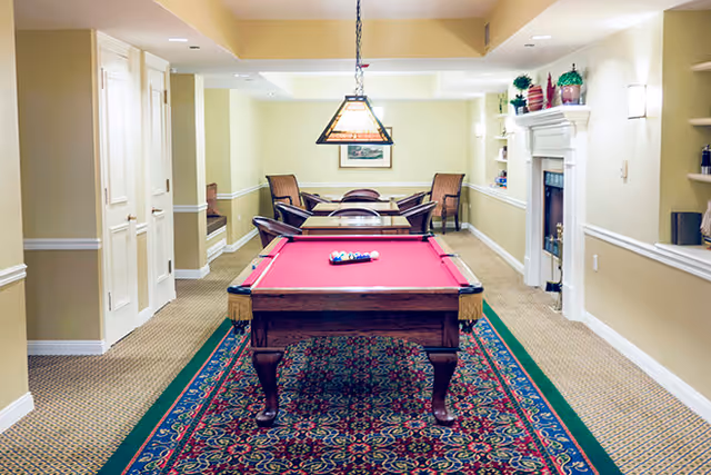 A well-lit recreational room featuring a red felt pool table centered on a patterned rug. Behind the pool table, there is a seating area with a table and four chairs. The room has beige walls with white trim, a fireplace on the right side decorated with plants and small items, and a hanging light fixture above the pool table.
