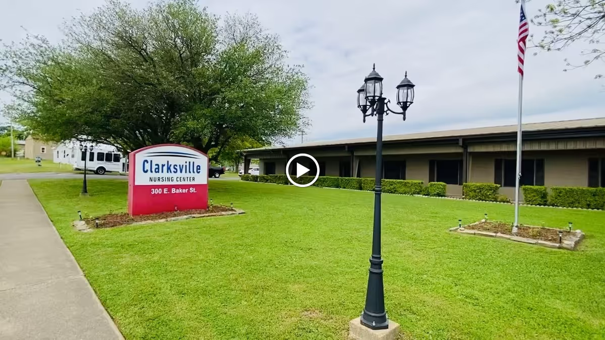 Exterior view of Clarksville Nursing Center showing a green lawn with a sidewalk, a street lamp, an American flag on a flagpole, and a sign with the facility's name and address. The building is a single-story structure with windows and bushes along the front.