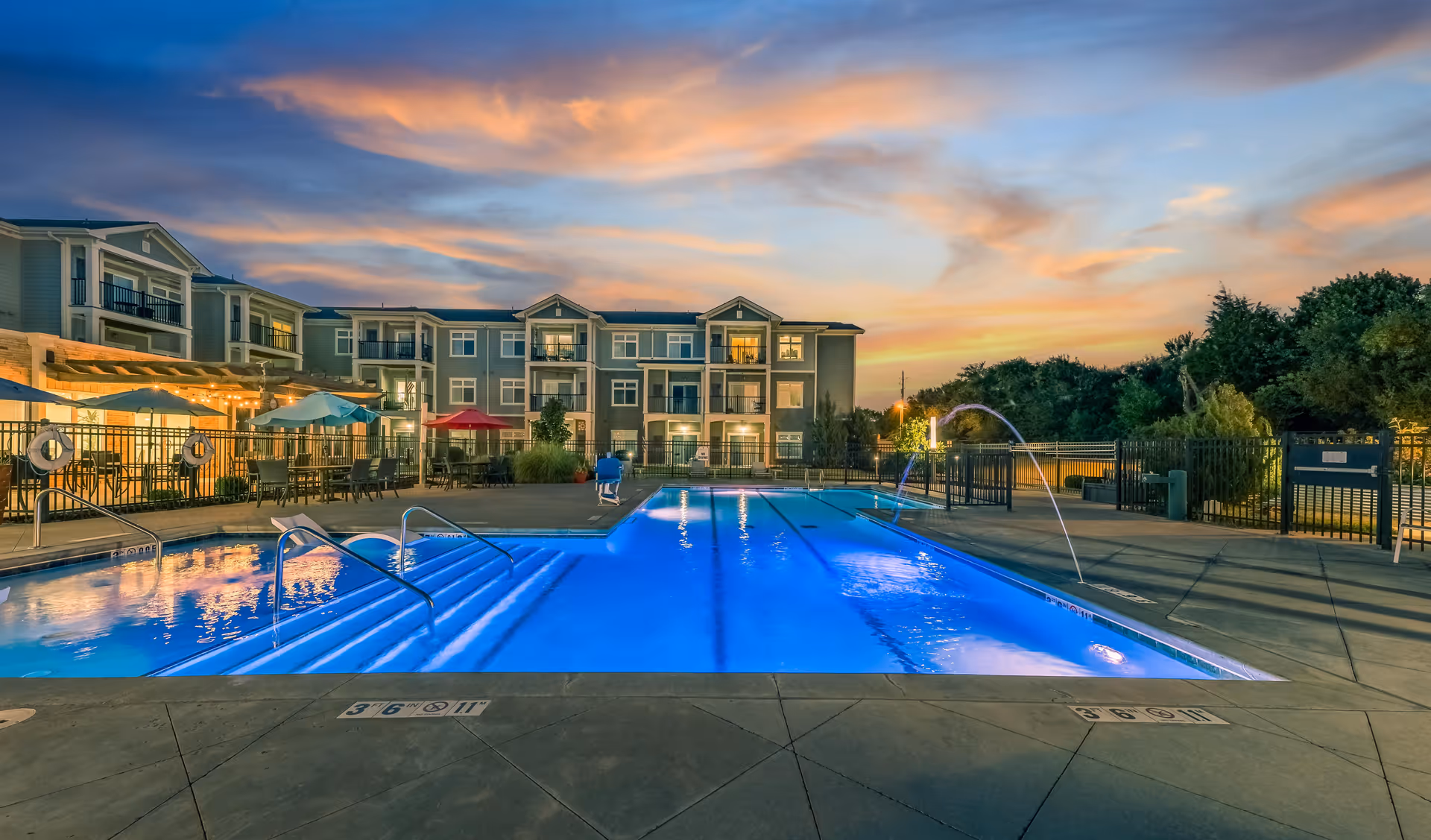 Outdoor swimming pool at dusk with blue water illuminated by pool lights, surrounded by a concrete deck with lounge chairs and umbrellas. In the background, there is a three-story residential building with balconies and warm interior lights, under a colorful sky with clouds at sunset.