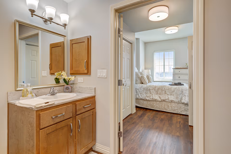 View of a bathroom vanity with a sink, mirror, and wooden cabinets on the left, leading into a bedroom with a bed, pillows, a window with blinds, and a dresser. The flooring is wooden and the walls are light-colored.