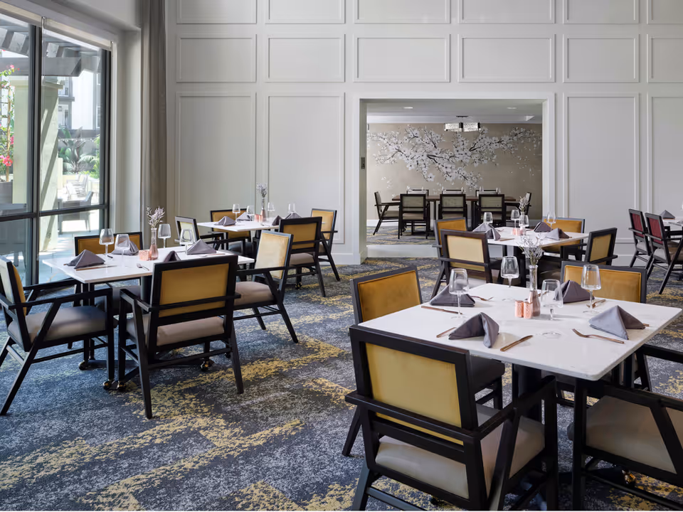 A bright dining room with multiple square tables set with wine glasses, napkins, and cutlery. The room features large windows on the left side allowing natural light to enter, beige and black chairs around the tables, and a patterned carpet. In the background, there is an adjoining room with a decorative wall featuring a floral mural and additional dining tables and chairs.