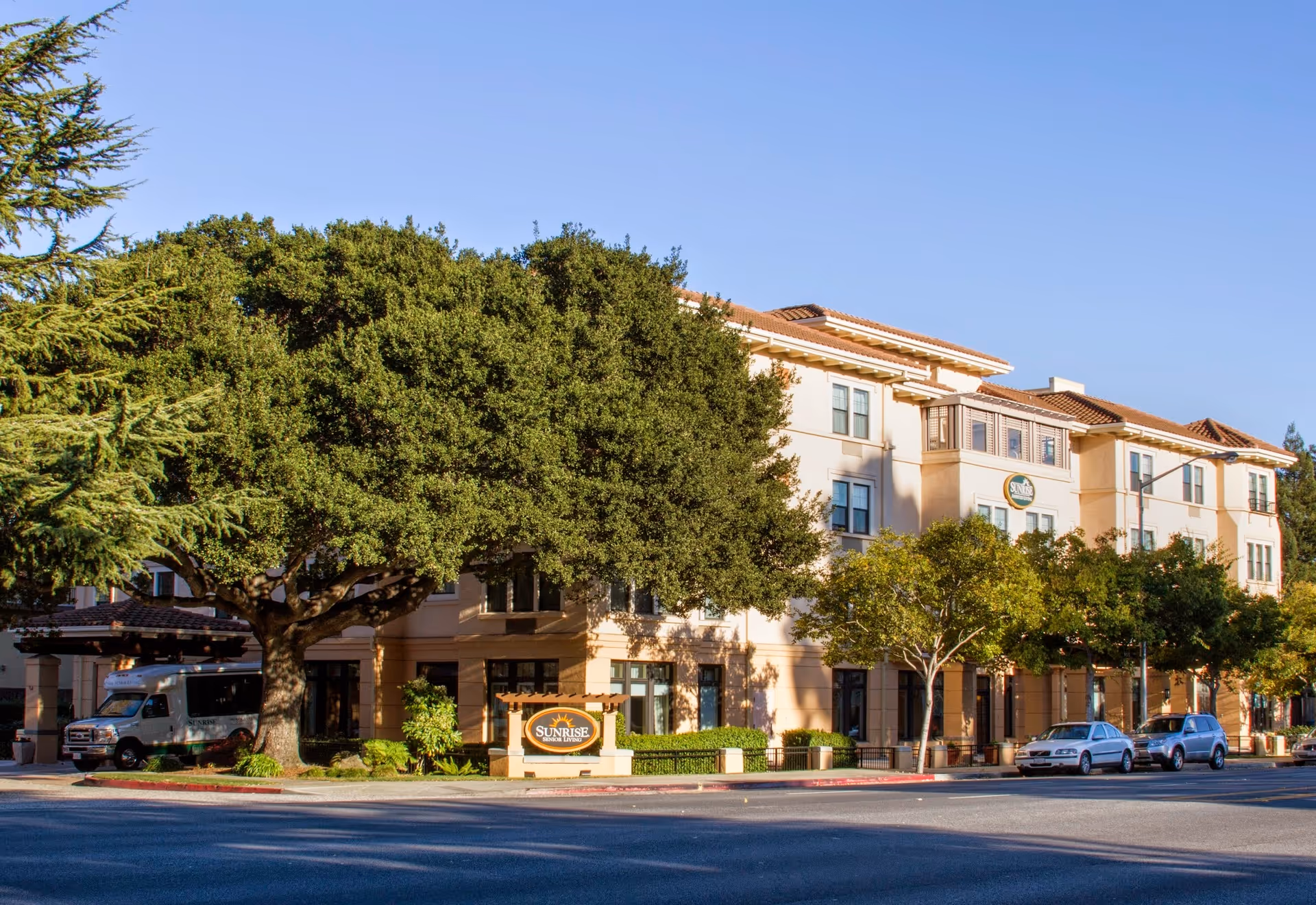 Exterior view of a multi-story senior living facility building with beige walls and a tiled roof, surrounded by large green trees and parked cars along the street. A sign in front reads 'Sunrise of San Mateo'.