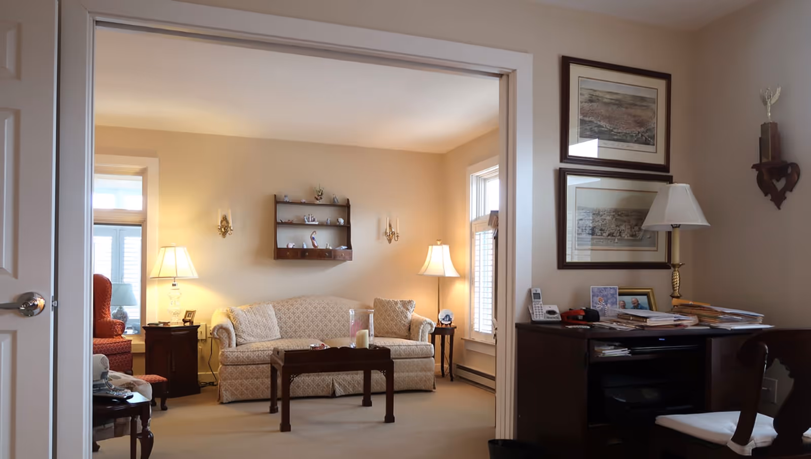 A cozy living room with a beige patterned sofa, two side tables each with a lamp, a wooden coffee table with candles, and a wall-mounted shelf with small decorative items. Adjacent to the living room is a small office area with a dark wooden desk cluttered with papers, a lamp, framed pictures, and two framed artworks hanging on the wall above the desk.