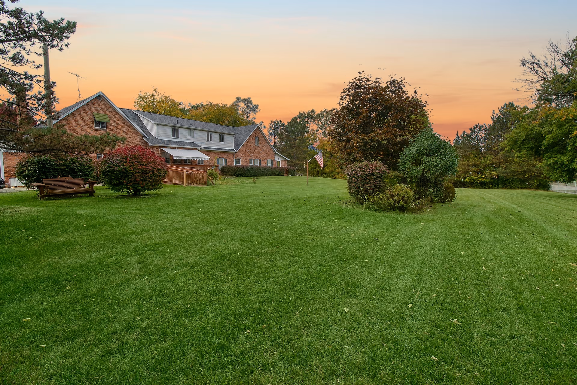 A large green lawn with neatly trimmed grass and several bushes and trees. In the background, there is a two-story brick building with multiple windows and a small porch. The sky is a warm orange, indicating either sunrise or sunset.